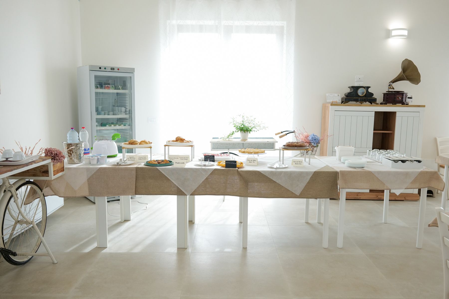 Una sala luminosa e bianca con un lungo tavolo apparecchiato per un buffet con vari dolci e bevande.