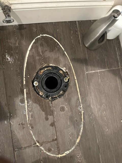 A toilet flange is sitting on a wooden floor in a bathroom.