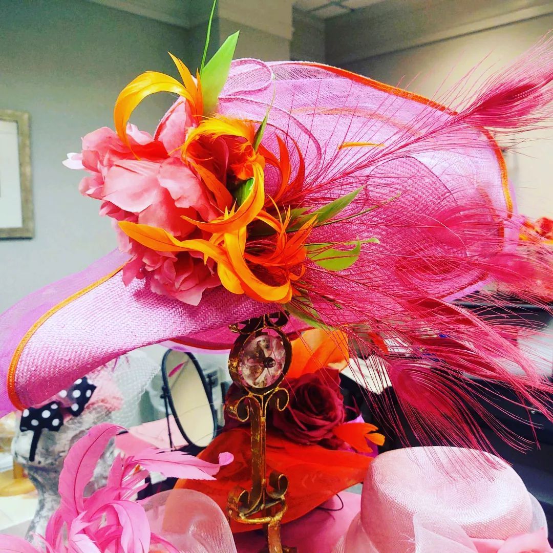 Pink wide-brimmed hat with orange and pink flowers, feathers, and embellishments, in a shop setting.