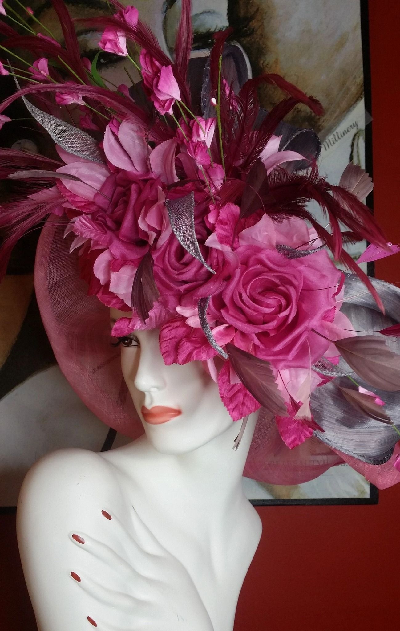 Mannequin wearing a large, elaborate pink and magenta hat with flowers and feathers. Red background.