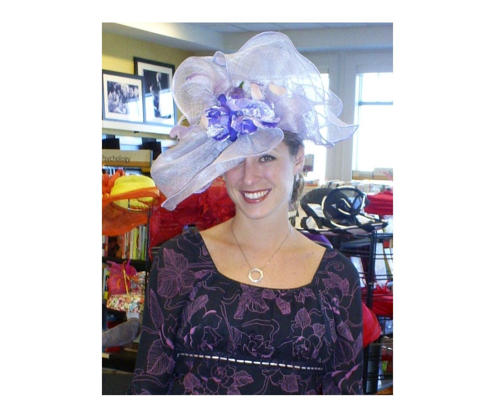 Woman wearing a large, lavender hat with purple flowers, smiling. She is in a store with hats in the background.