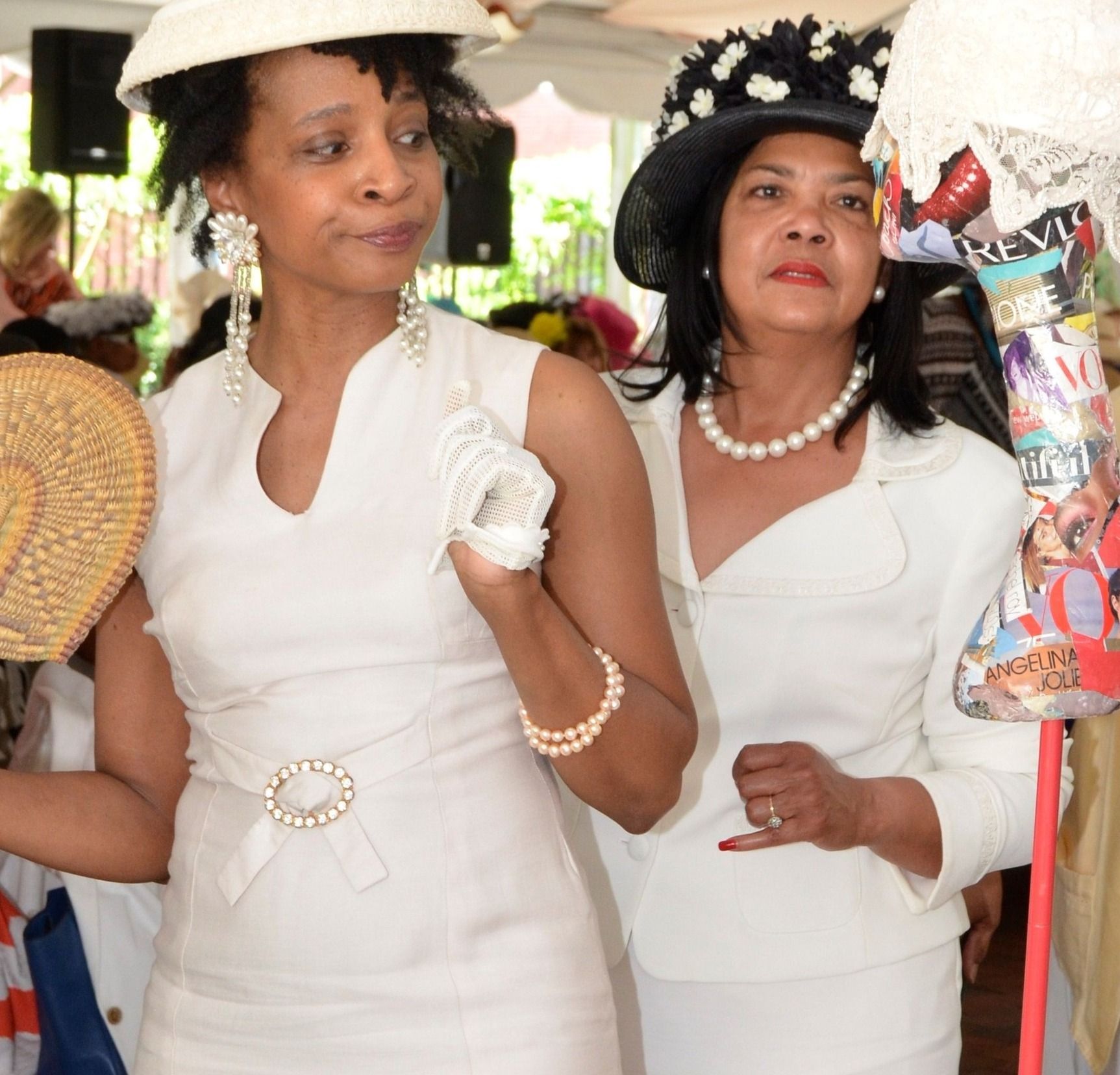 Two women in white outfits, wearing hats, looking at hats for sale.
