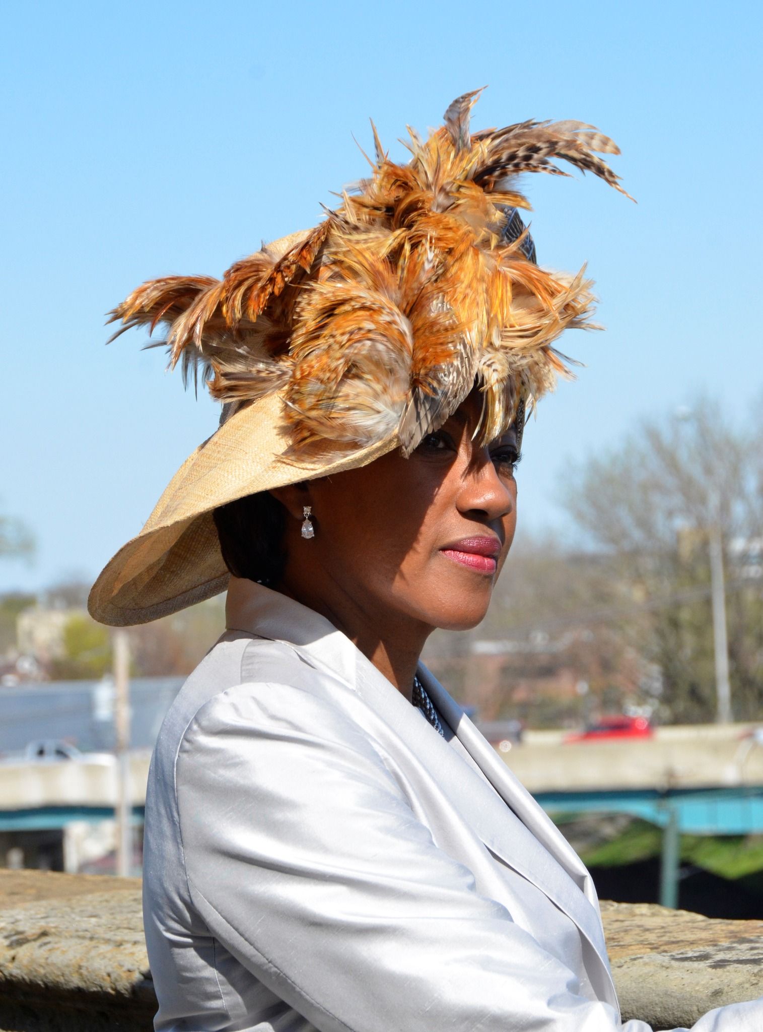 Woman in straw hat with brown feathers, wearing a silver jacket, outdoors on a sunny day.