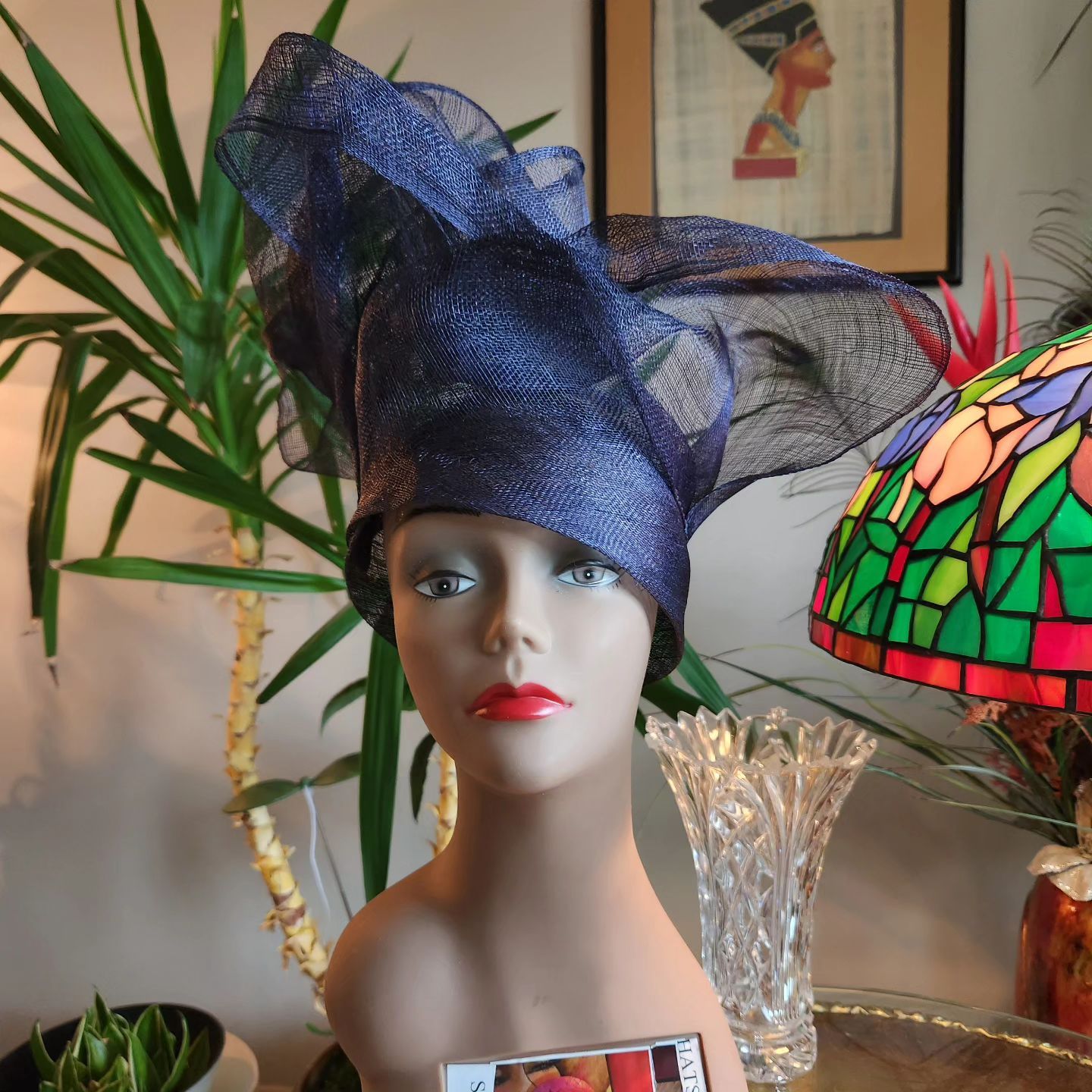 Mannequin head wearing a navy blue fascinator, in a room with plants and a stained glass lamp.