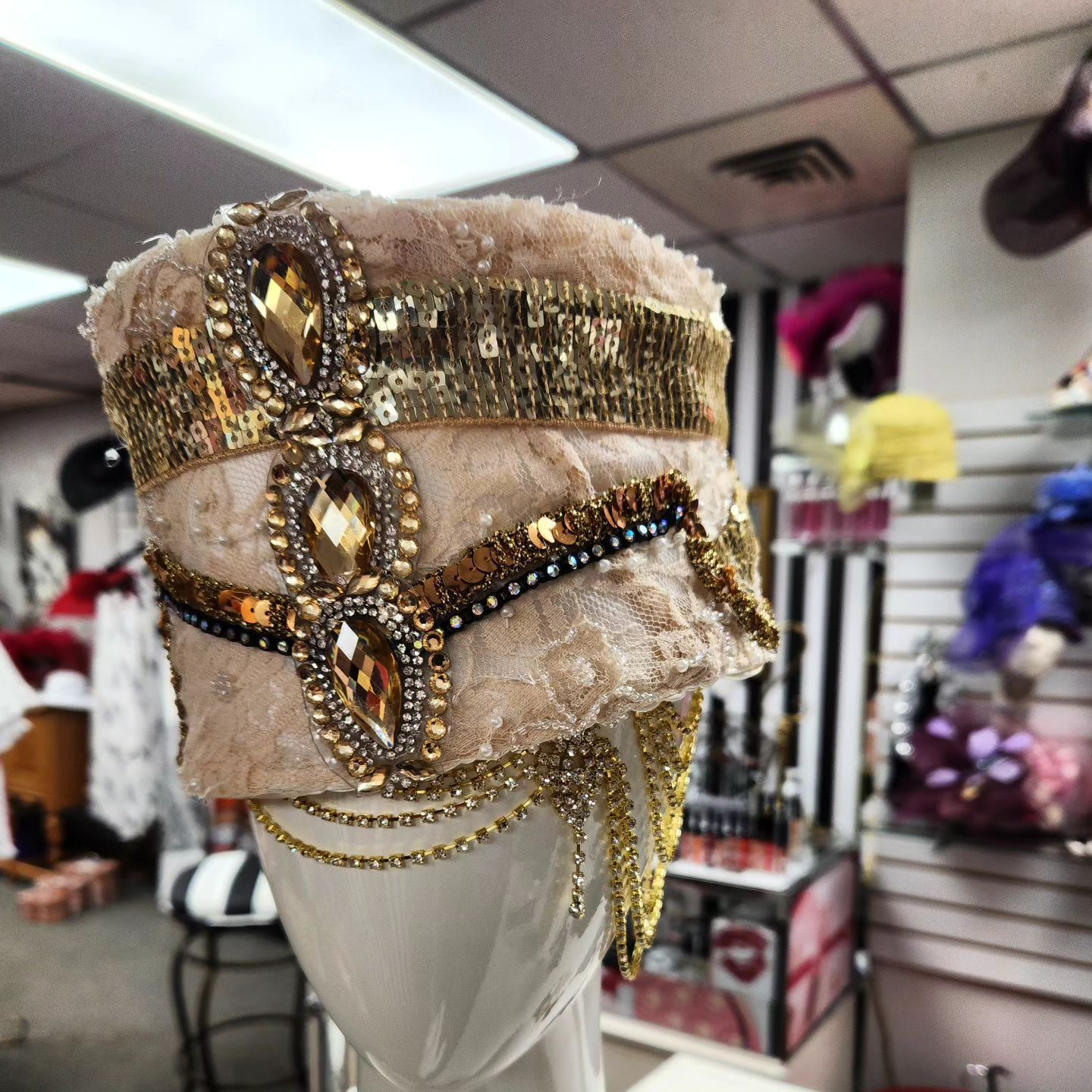 Ornate, bejeweled beige and gold hat on a white mannequin head in a shop setting.
