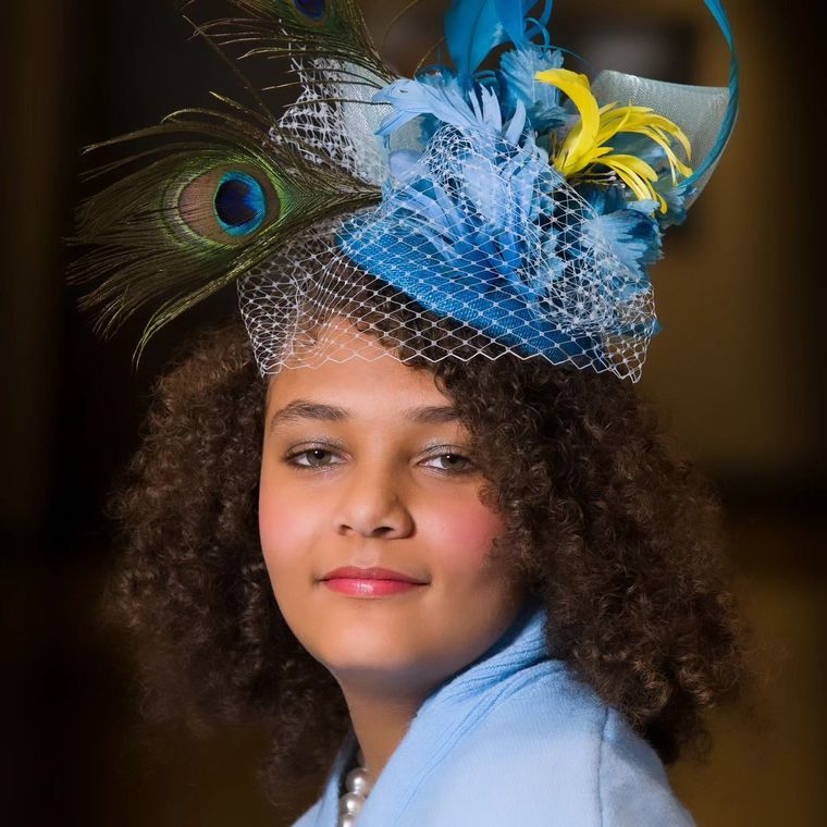 Woman wearing a blue hat with peacock feathers, tulle, and blue and yellow feathers, smiling.
