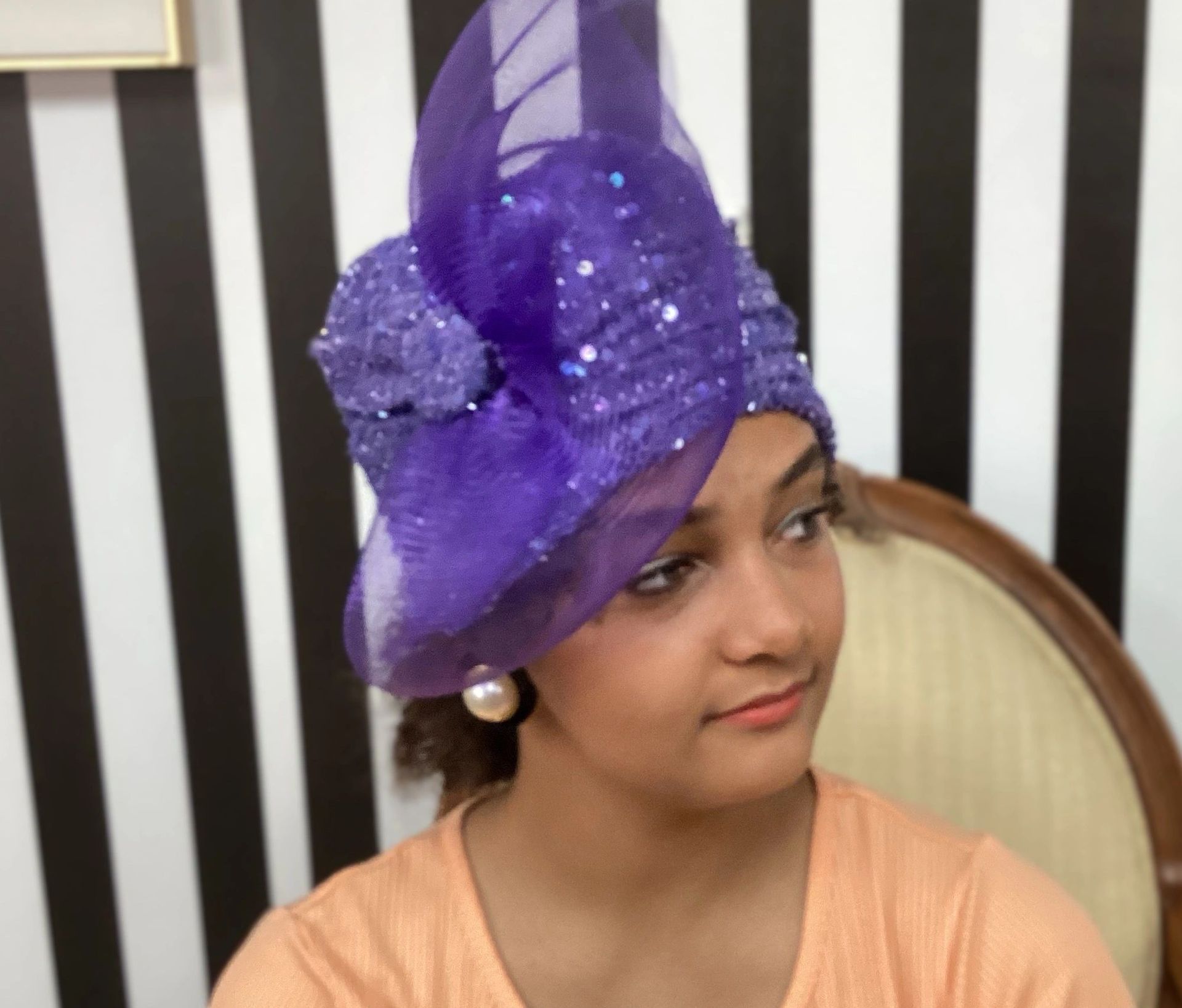 Woman wearing a purple, sequined headpiece with a large pearl earring. She is seated in front of a striped wall.