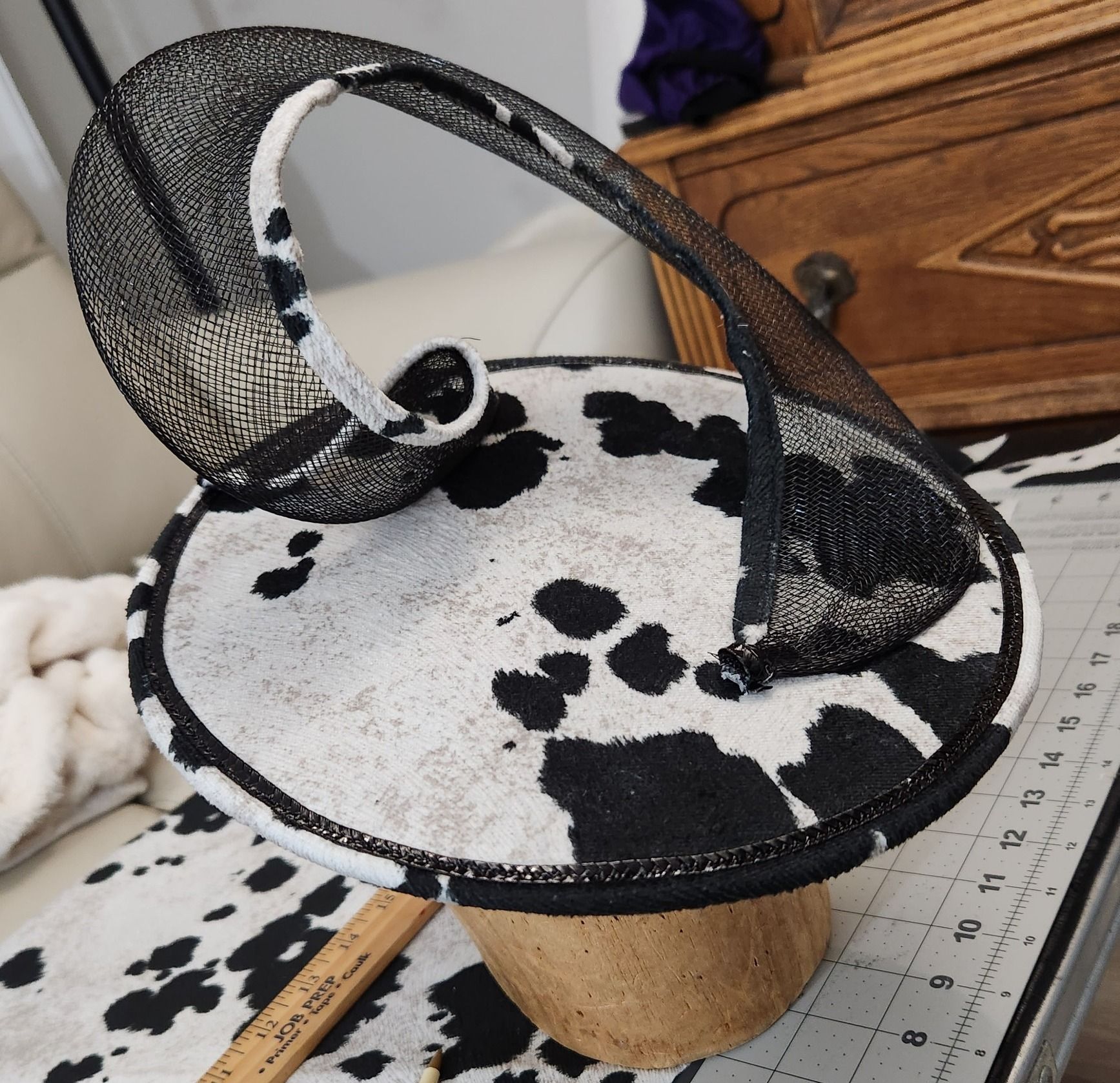 A black and white cow-print hat with a woven black brim, sitting on a wooden stand.