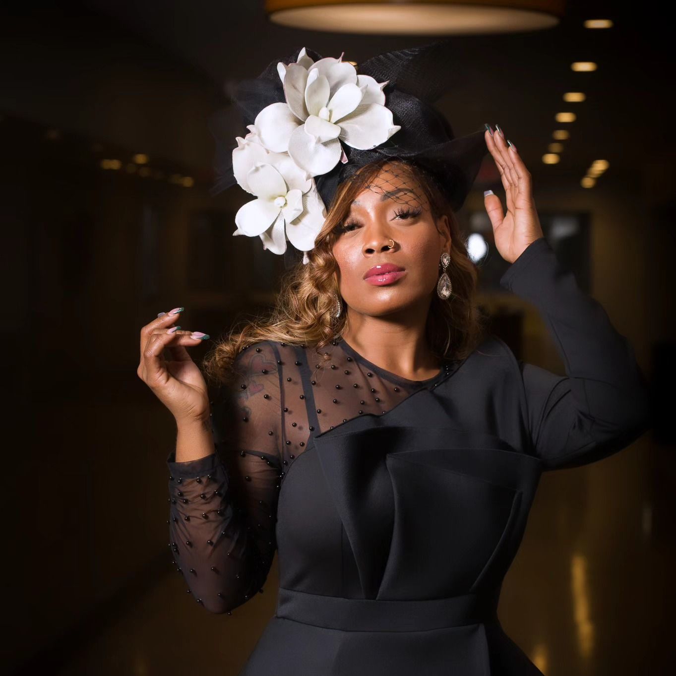 Woman in black dress and floral headpiece, raising hands, indoors with lighting.