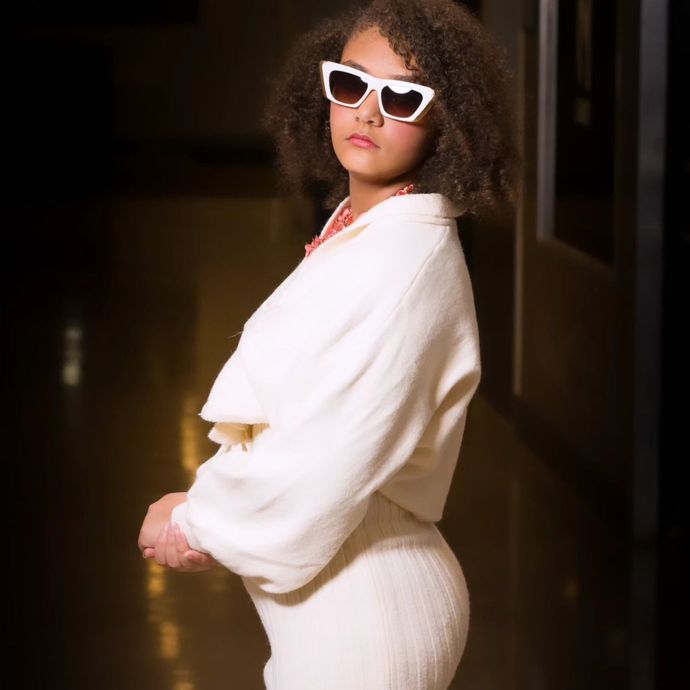 Person in white outfit and heart-shaped sunglasses, standing in a hallway, posing.