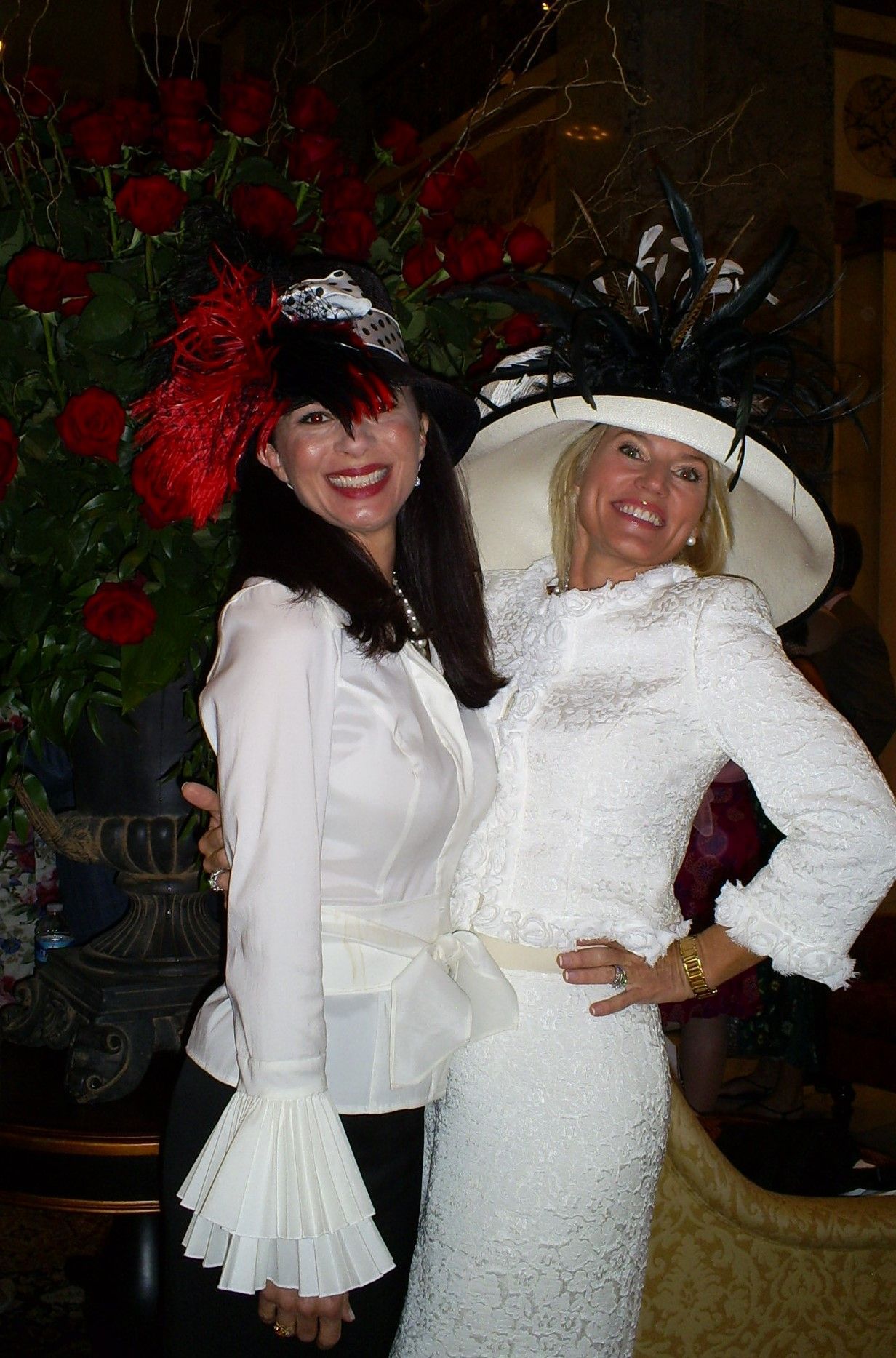 Two women in hats pose together, one wearing white, the other black. Red roses in background.