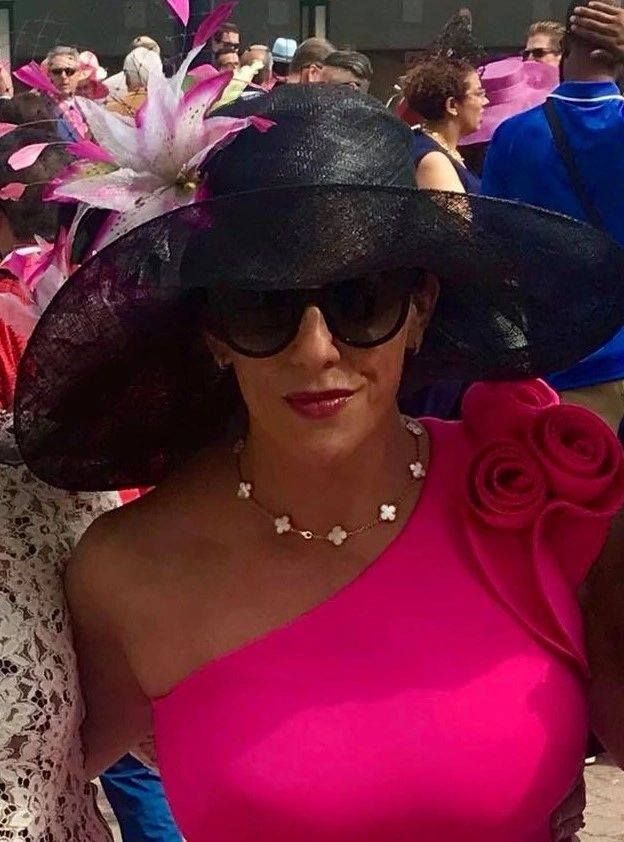 Woman in large black hat and pink dress, wearing sunglasses, at an outdoor event.