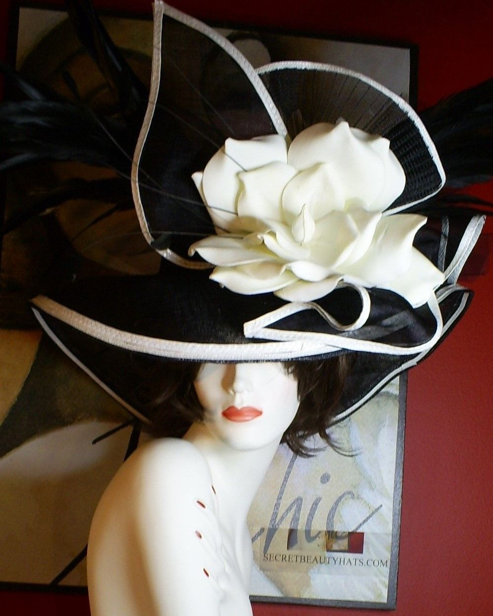 Mannequin wearing a large black and white floral hat with a white flower. Red background.
