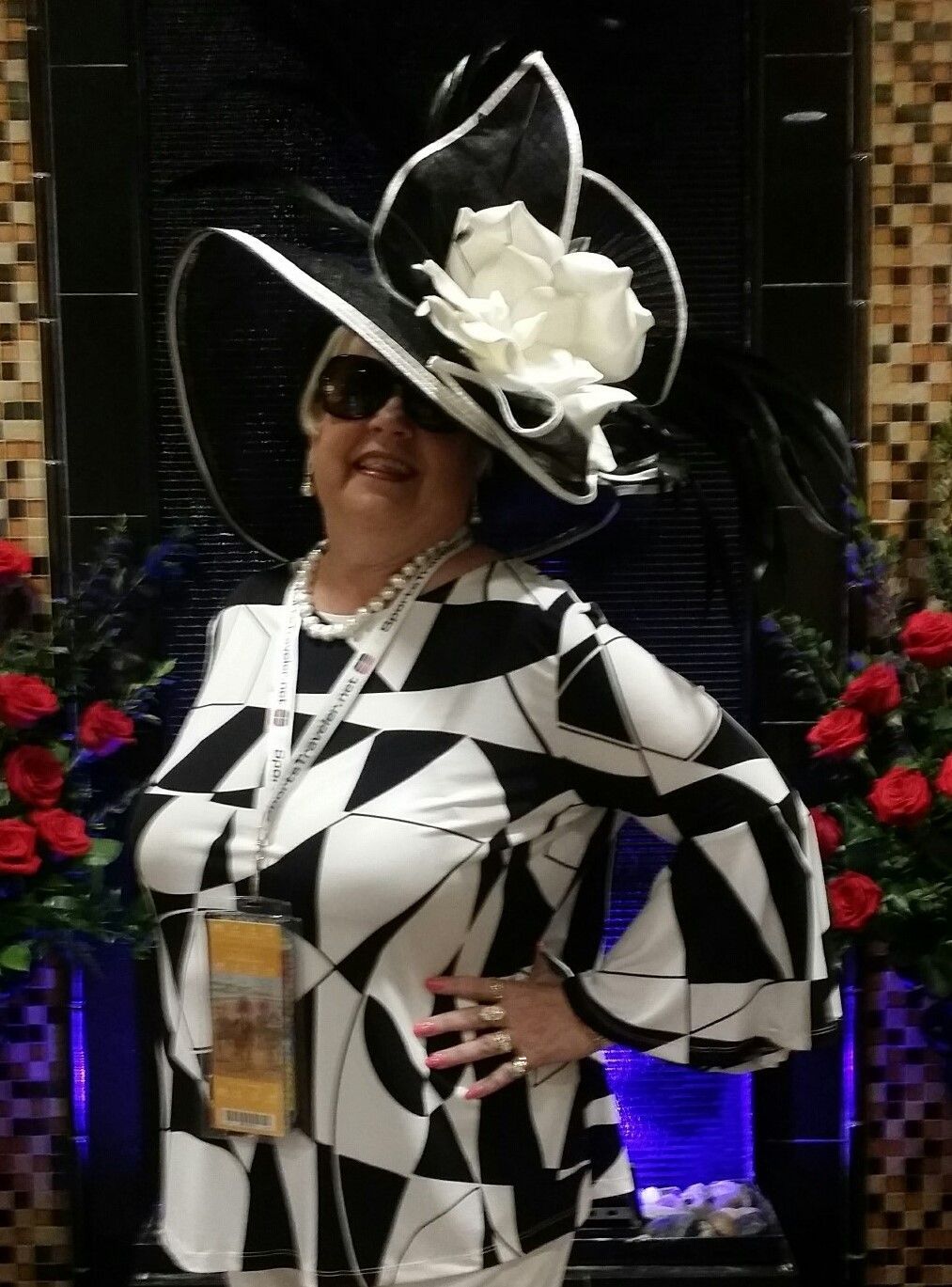 Woman in a large black and white hat and geometric print outfit, standing in front of a fountain.