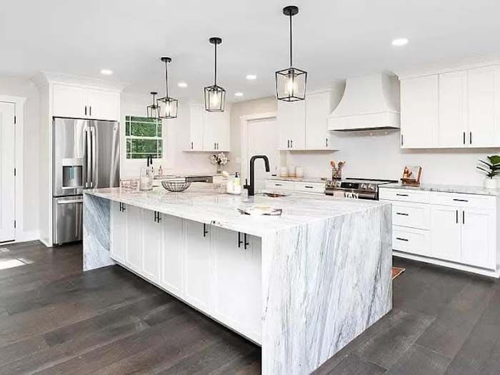 Modern white kitchen with marble island, dark wood floors, stainless steel appliances, and pendant lights.