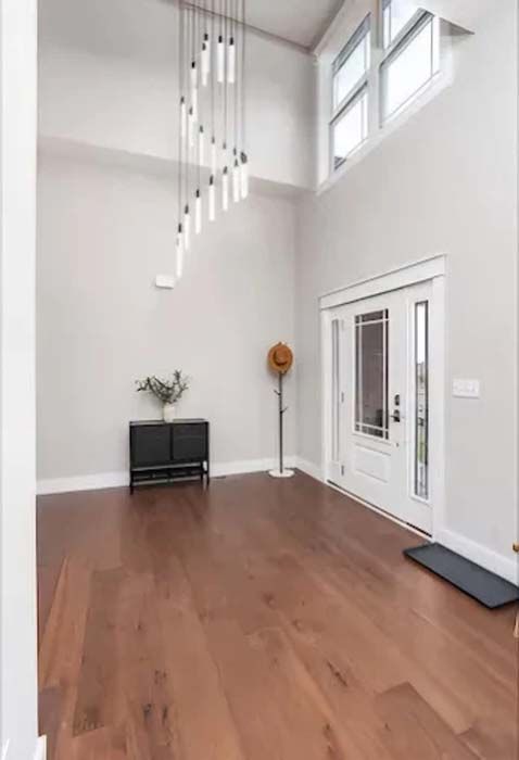 Spacious entry hall with wood floors, high ceiling, modern light fixture, black cabinet, and white door.