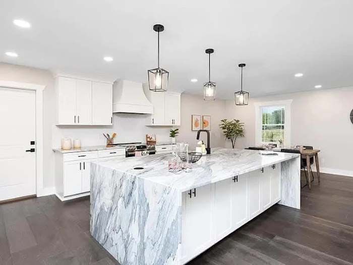 Modern white kitchen with marble island, dark wood floors, pendant lights, and stainless steel appliances.