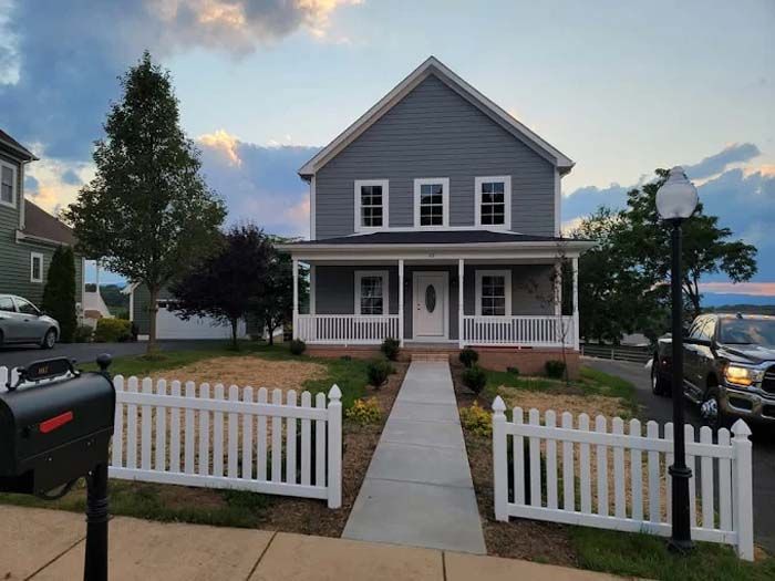 Gray house with white porch and picket fence, sidewalk leading to front door, cars parked nearby.