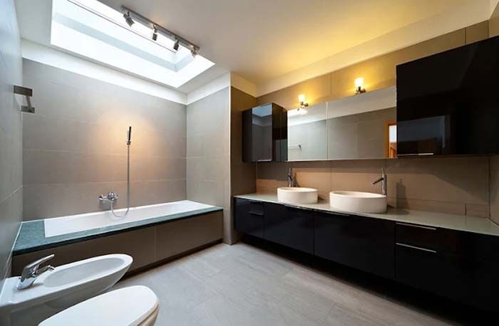 Modern bathroom with bathtub, double sinks, toilet, and black cabinets. Skylight illuminates the room.