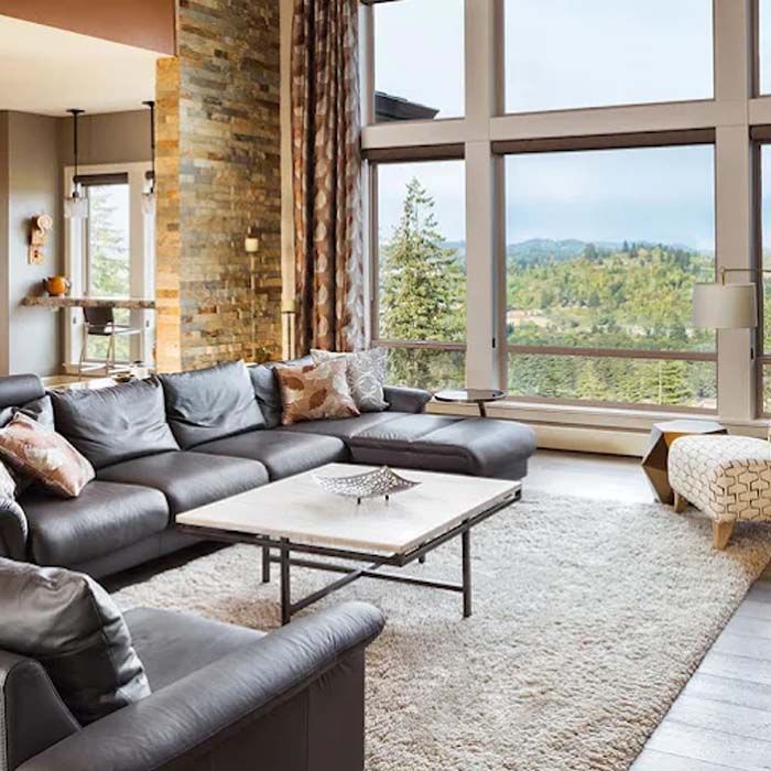 Living room with gray sectional sofa, large windows overlooking trees, and stone wall.