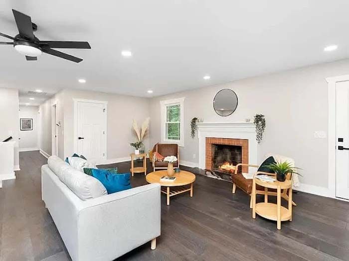Living room with fireplace, white walls, dark wood floor, sofa, chairs, round coffee table, and ceiling fan.