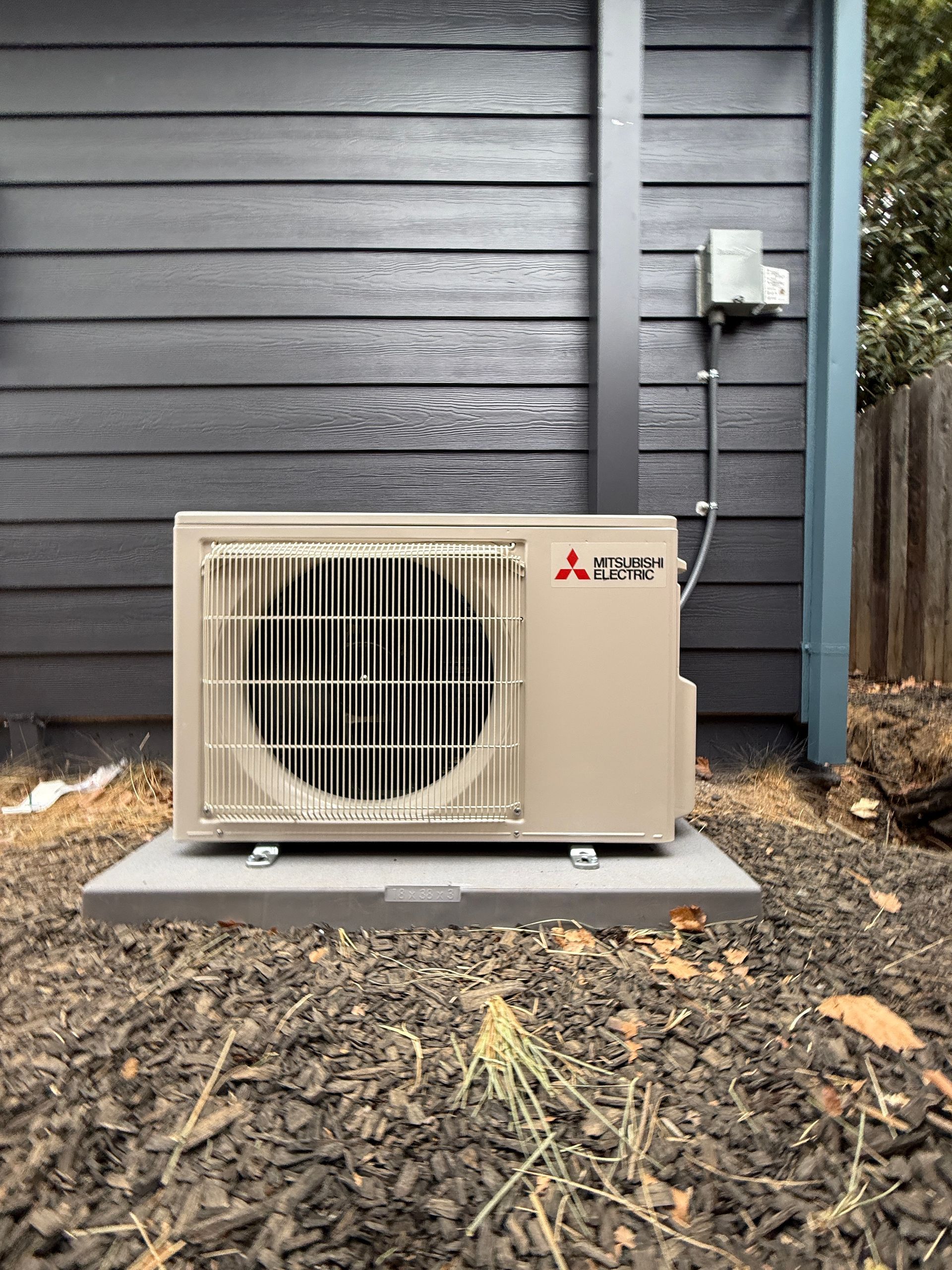 Outdoor Mitsubishi mini-split condenser beside a dark gray house wall on a concrete pad.