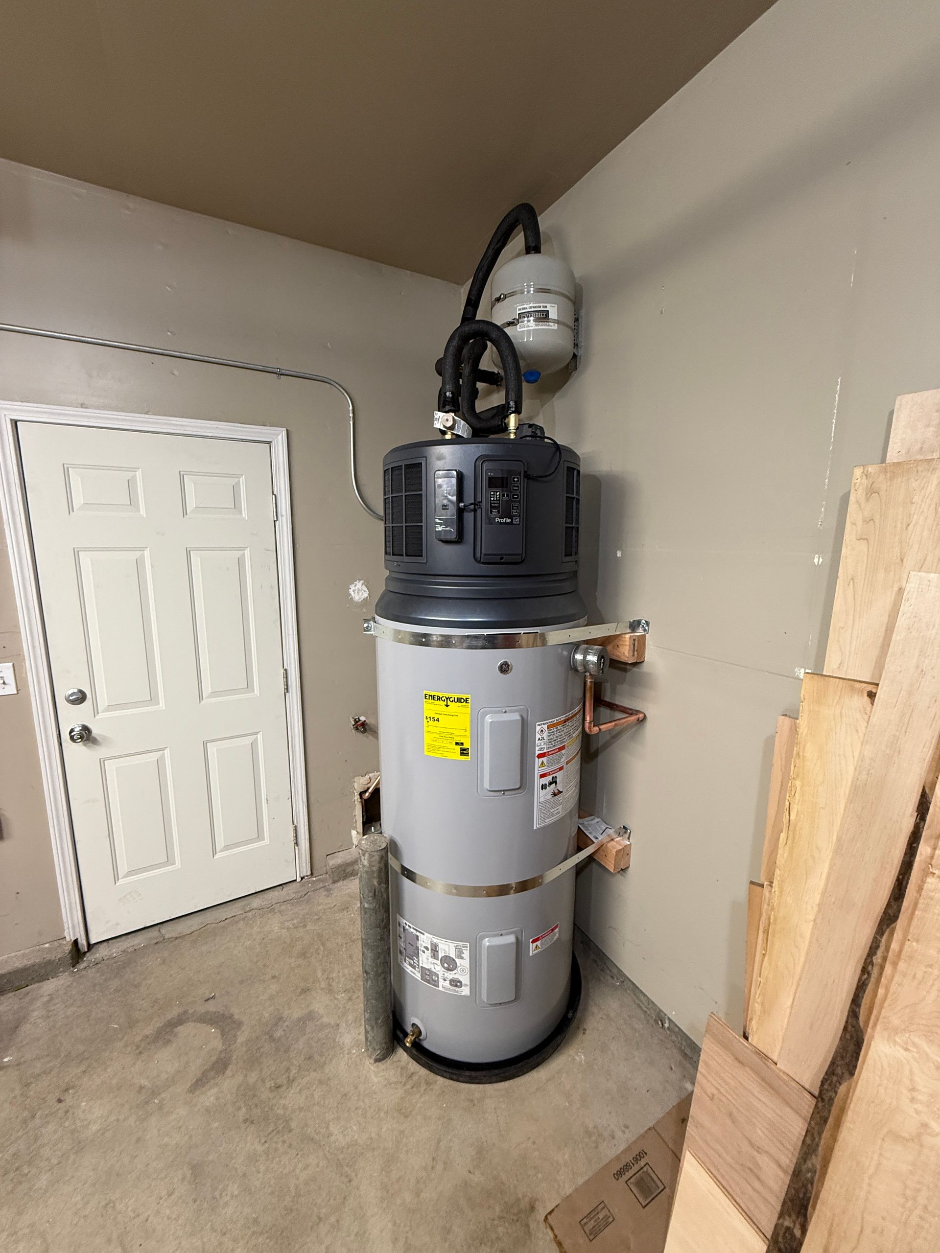 Tall gray water heater tank in a utility room with stacked wood and a white door nearby