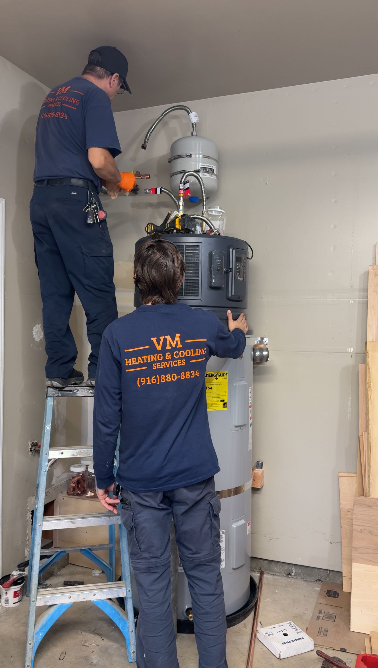 Two technicians in blue uniforms work on a large tank in a room, one on a ladder, one at the base.