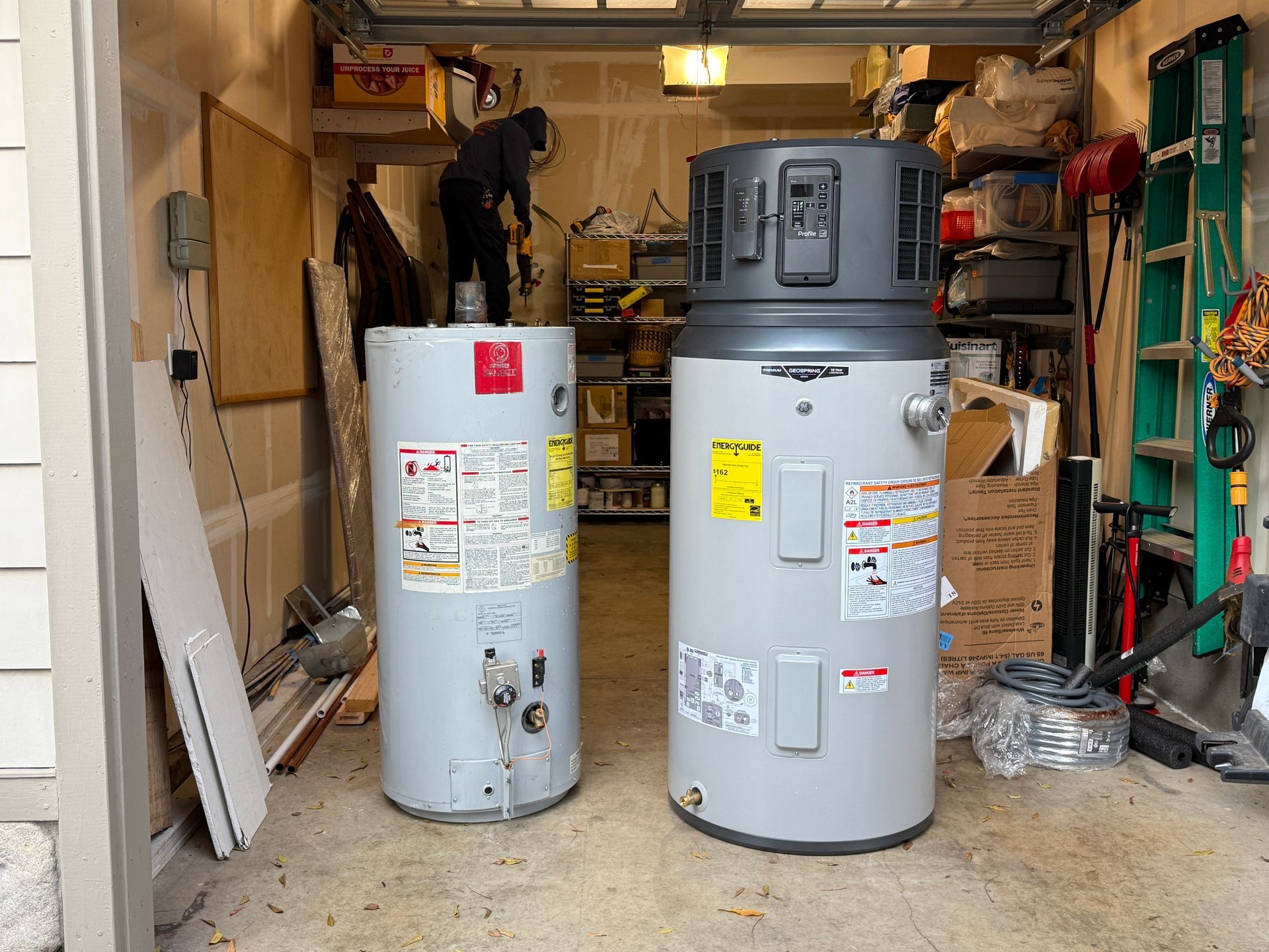 Two water heaters in a cluttered garage workshop, one tall gray and one shorter white.