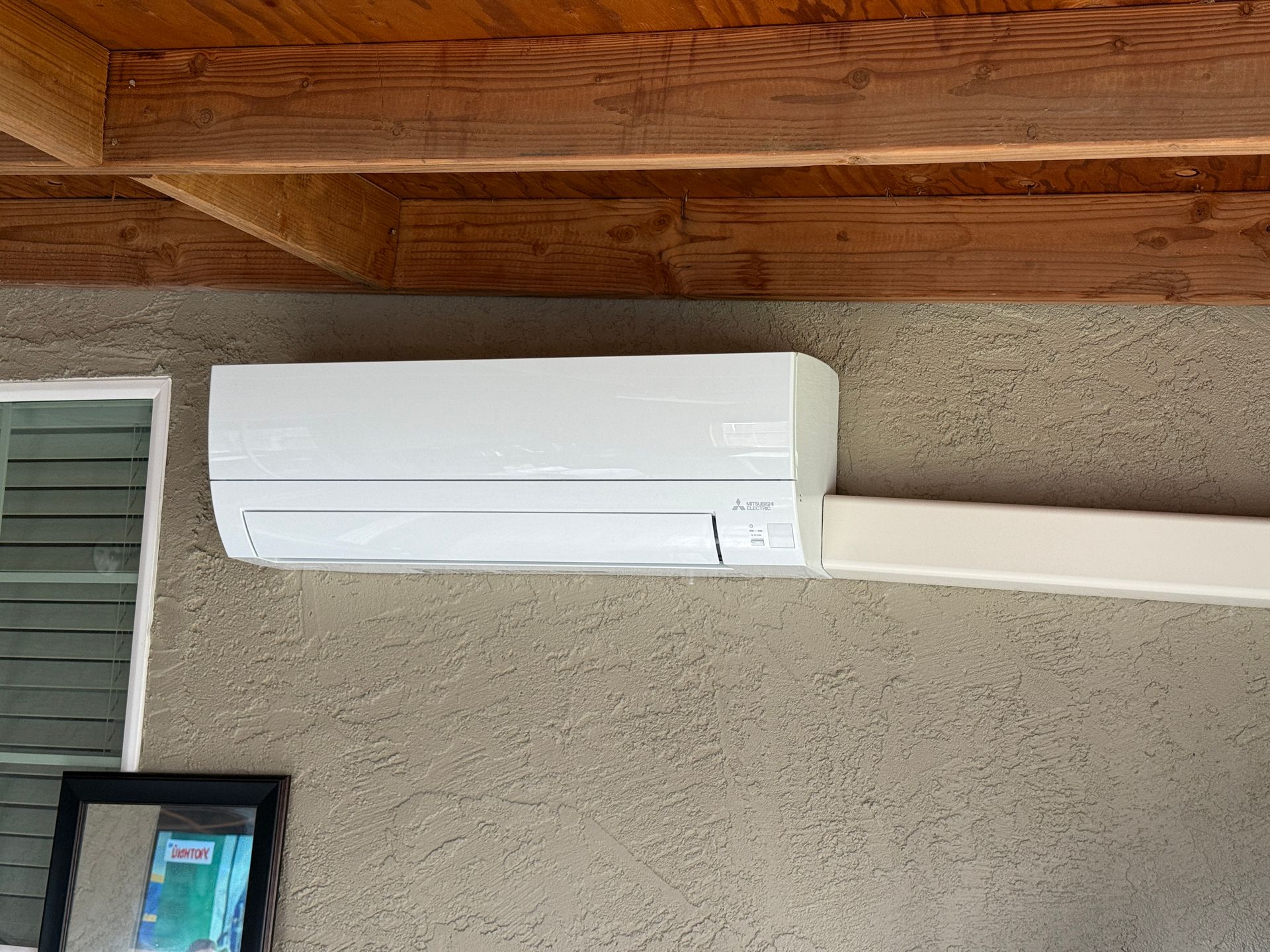 White wall-mounted air conditioner under a wooden patio roof, with a conduit running right.