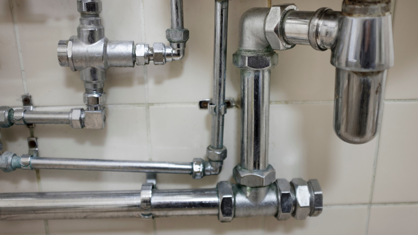 Shiny silver pipes and valves mounted on a beige tiled wall, with a cylindrical filter on the right.