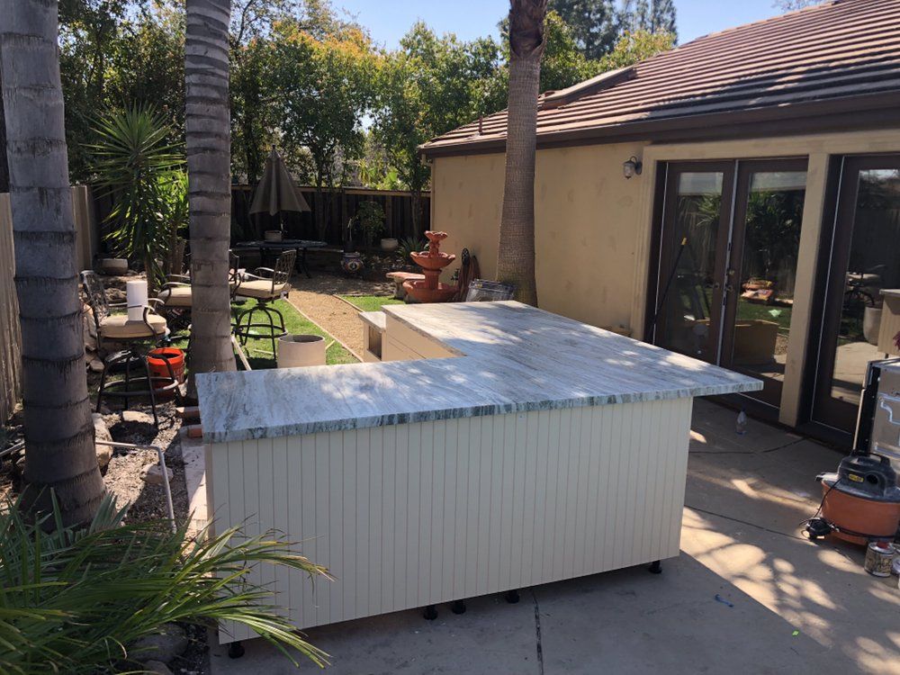 Outdoor kitchen with a white countertop and base, on wheels, next to a house and palm trees.