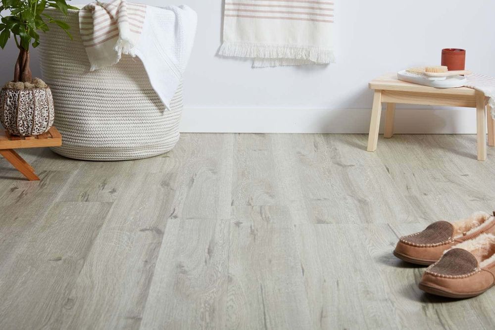 Bathroom scene with light wood-look flooring, basket, towels, small stool, slippers, and decor.