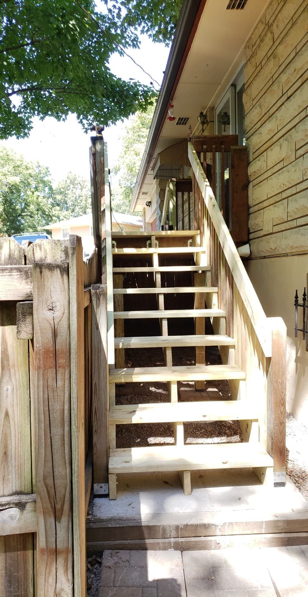 Wooden outdoor stairs leading to a house. Light brown wood, fence on left, green trees overhead.