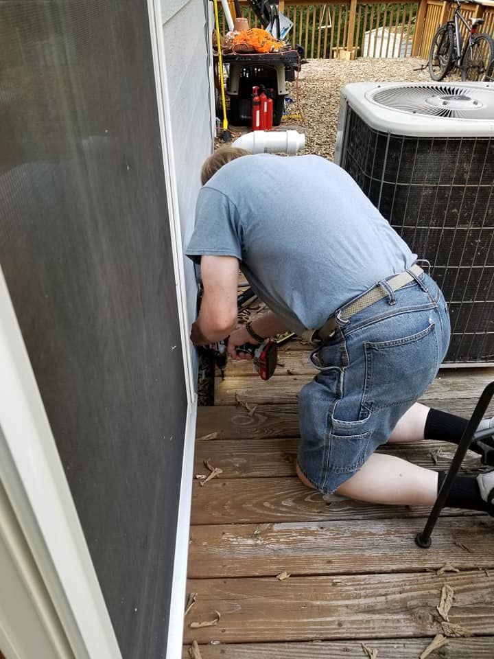 Man kneeling on deck, using a drill to work near a screen door and AC unit.
