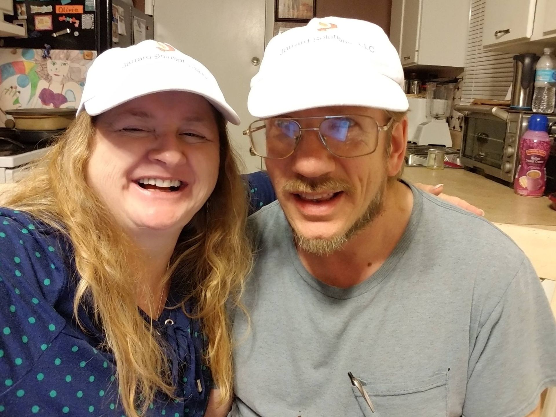 Two people, a woman and a man, wearing white caps, smiling in a kitchen.