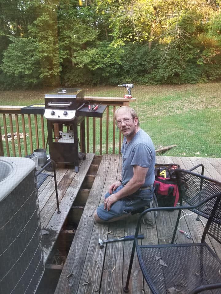Man kneeling on deck, working near grill. Green trees in background, sunlight.