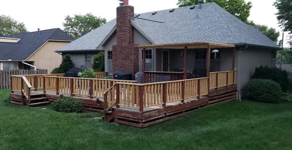 Wooden deck with railings and a pergola extending from the back of a house with a brick chimney.
