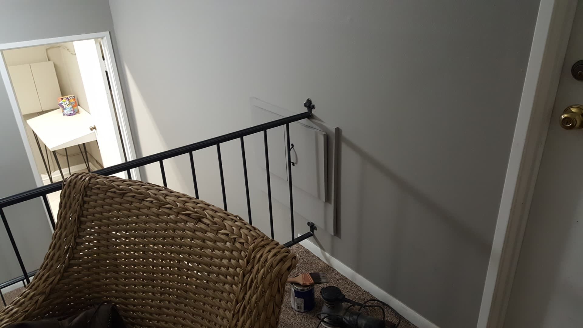 Wicker chair by a black railing, overlooking a hallway with a small, white room. Gray walls.