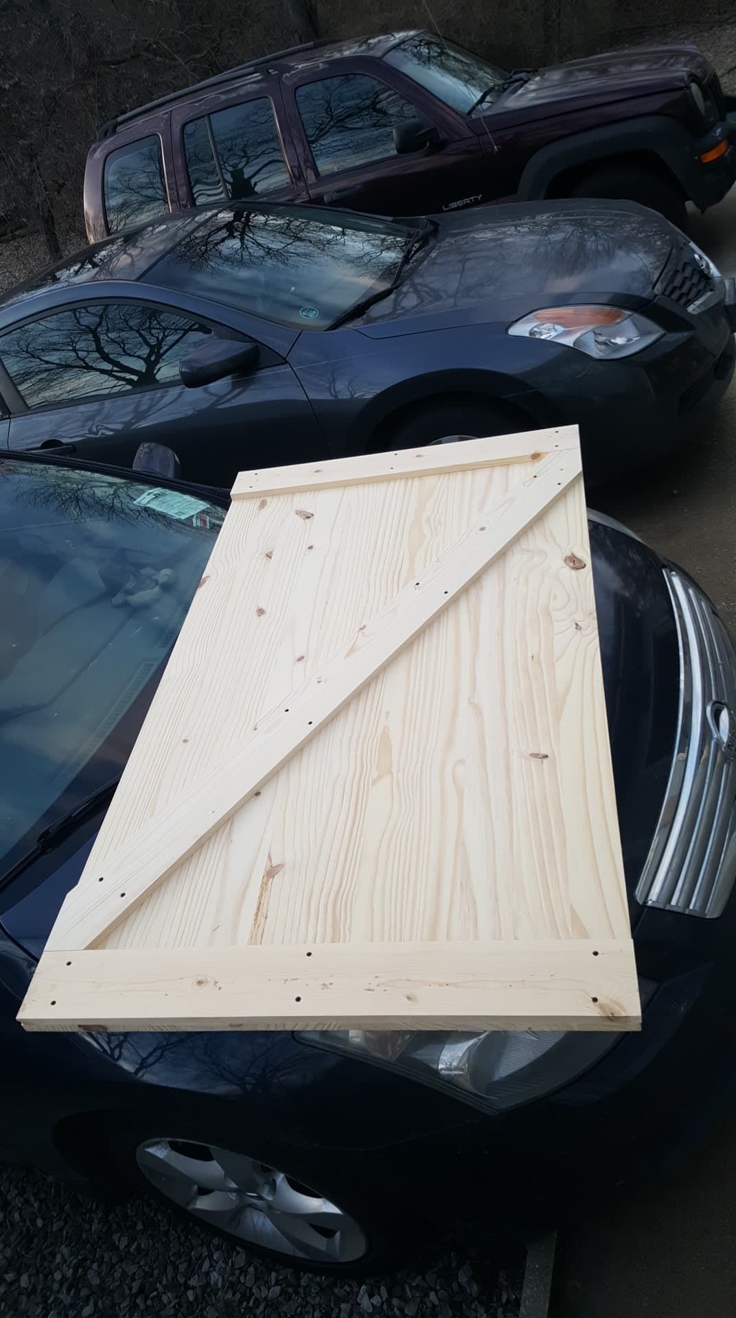 A wooden plank, with a diagonal brace, rests on the hood of a dark car, other cars in the background.
