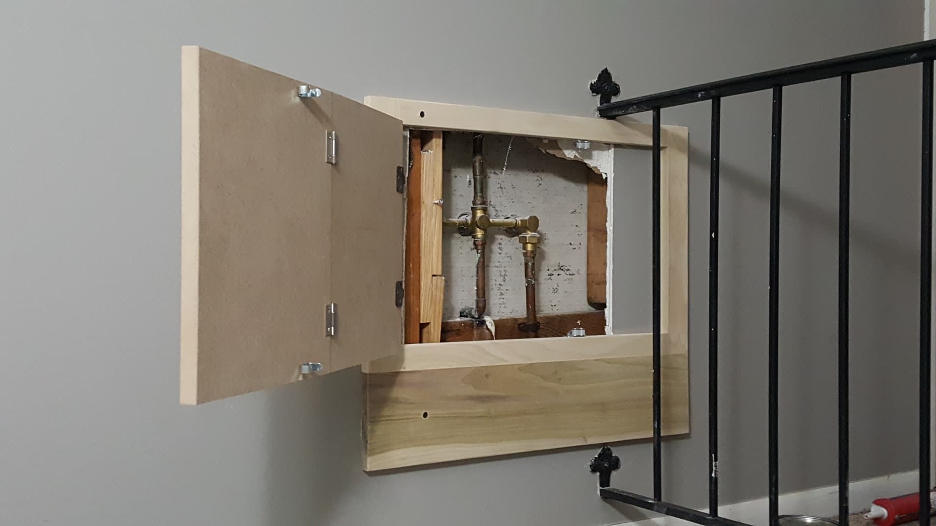 A plywood access panel on a gray wall, revealing plumbing. The panel is open, near a black railing.