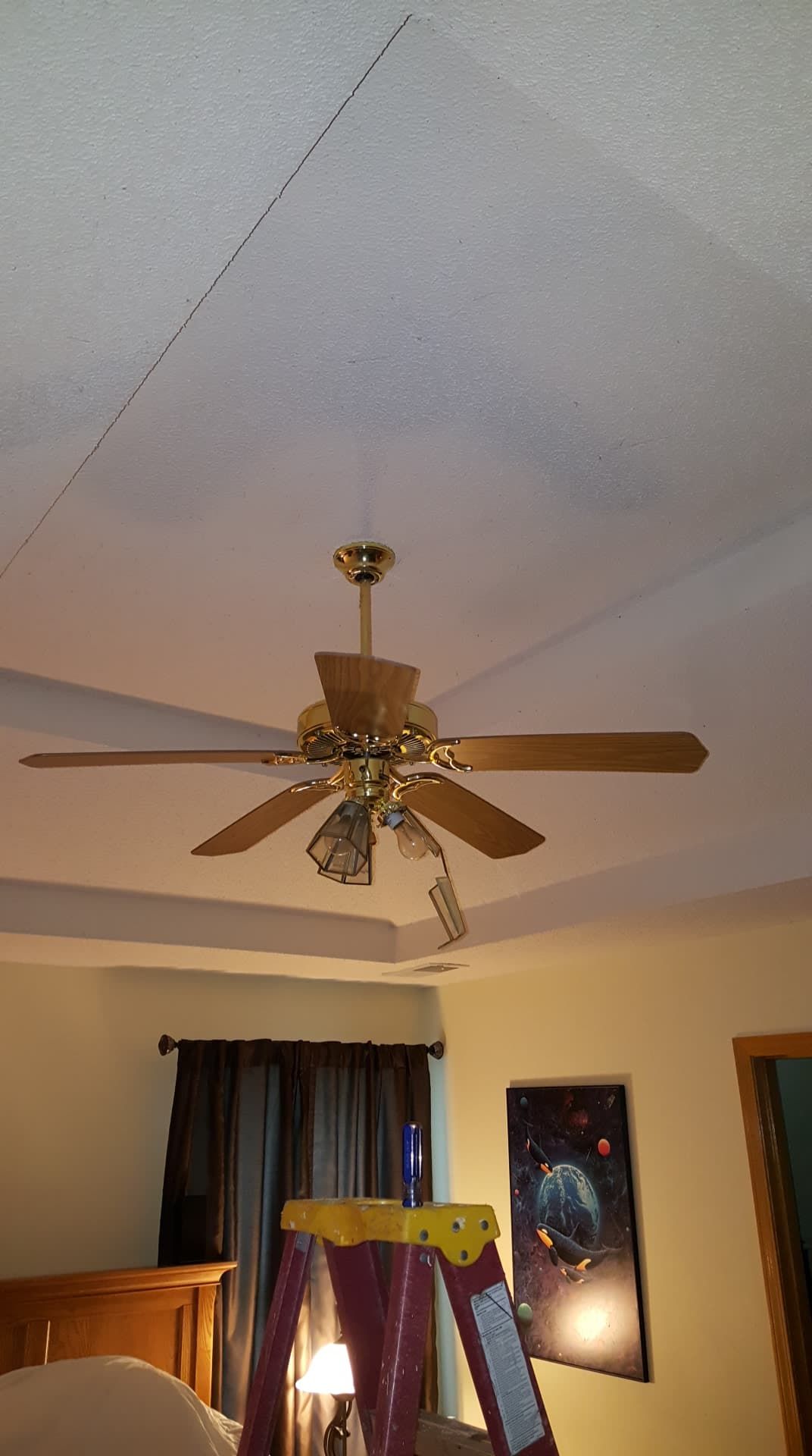 Ceiling fan below a cracked ceiling with a ladder. A framed art piece and a wooden bed are in the room.
