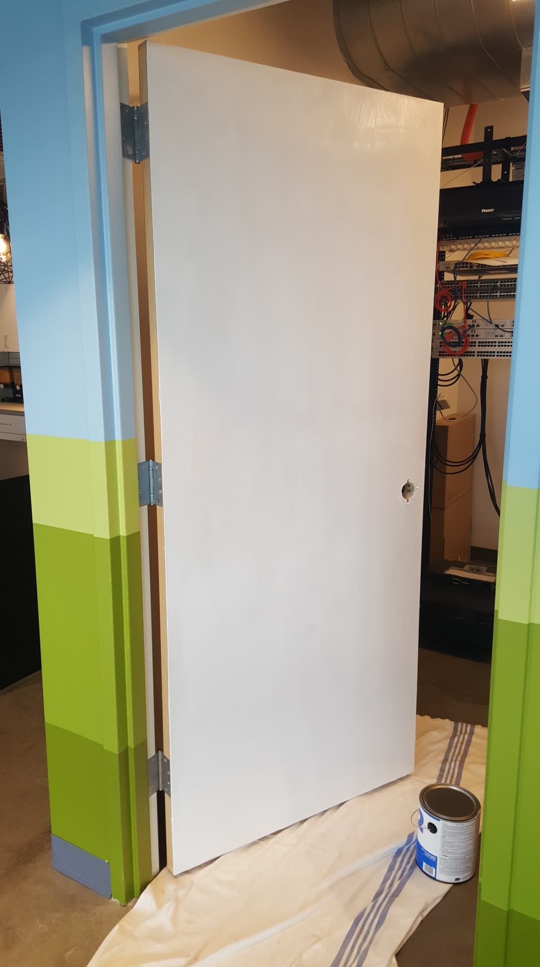 White painted door ajar in a basement. Walls are blue, green, and yellow. A paint can sits on the floor.