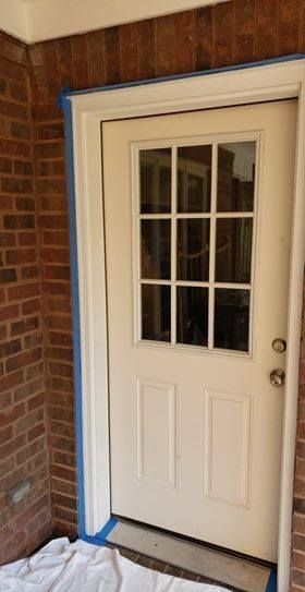 White door with window panes being painted, blue tape along brick wall, outdoor setting.
