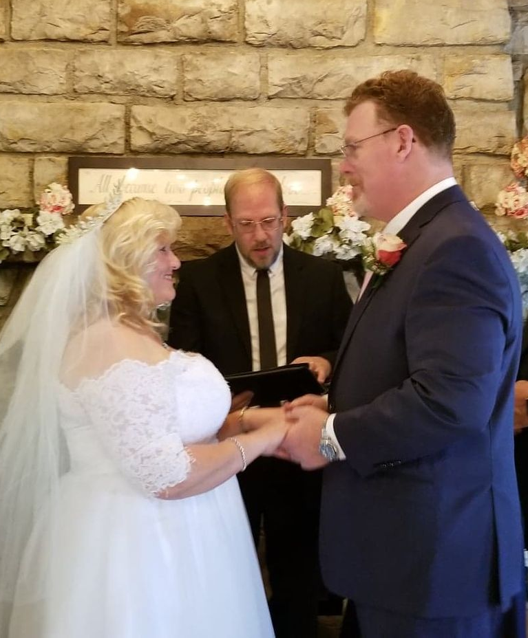 Bride and groom holding hands during wedding ceremony, officiant present. Stone wall and floral decorations.