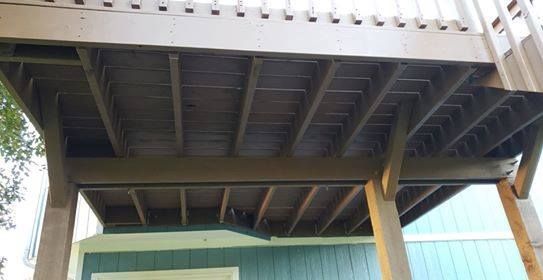 Underside view of a wooden deck with support beams; brown and gray tones, built over a light green house wall.