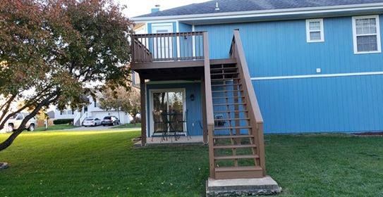 Two-story wooden deck with stairs attached to a blue house, overlooking a grassy yard.