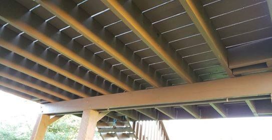 View from under a brown deck showing joists, support beams, and deck boards in a grid-like pattern.