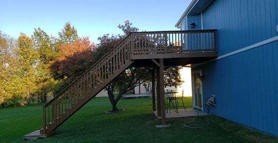 Wooden deck and stairs attached to a blue house, with green grass and trees in the background.