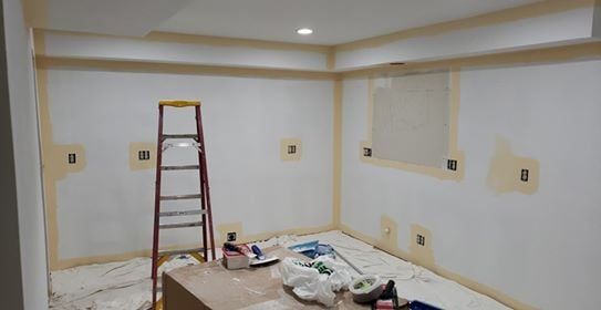 Room being painted, with ladder, masking tape, and supplies on the floor. White walls, yellow trim.